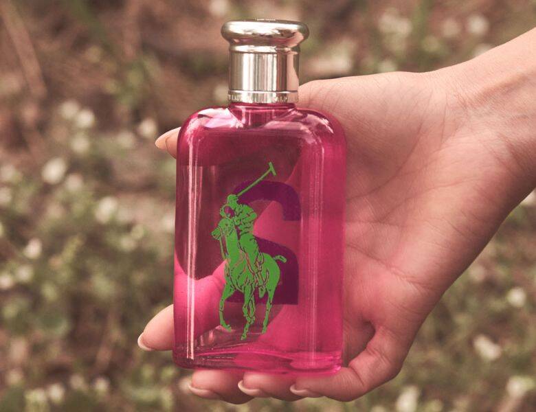 Her ses et billede af en parfumeflakon fra Polo Ralph Lauren holdt i en hånd. Flakonen er i en pink nuance med det grønne polo-logo på fronten og har et sølvfarvet låg. Baggrunden er sløret med naturprægede farver.