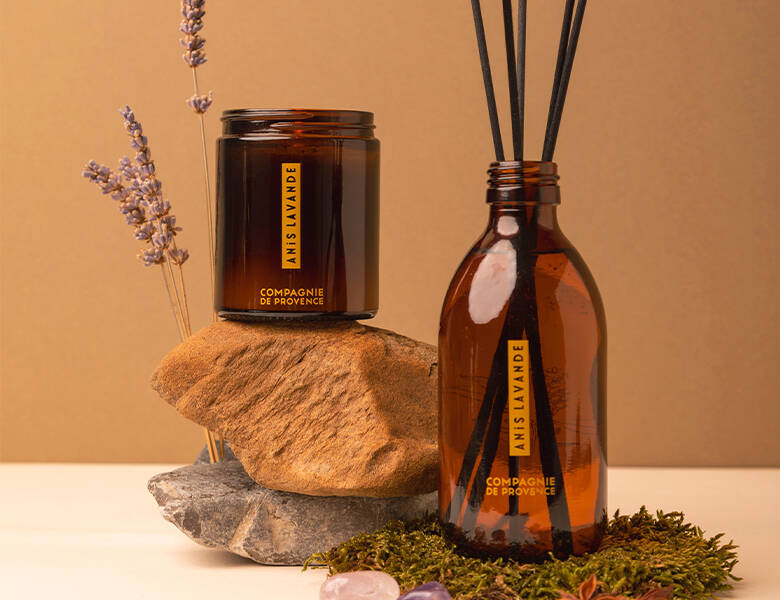 Her ses en brun baggrund med noget mos, sten og blomster. I midten ses to brune produkter - et duftlys og nogle duftpinde. 