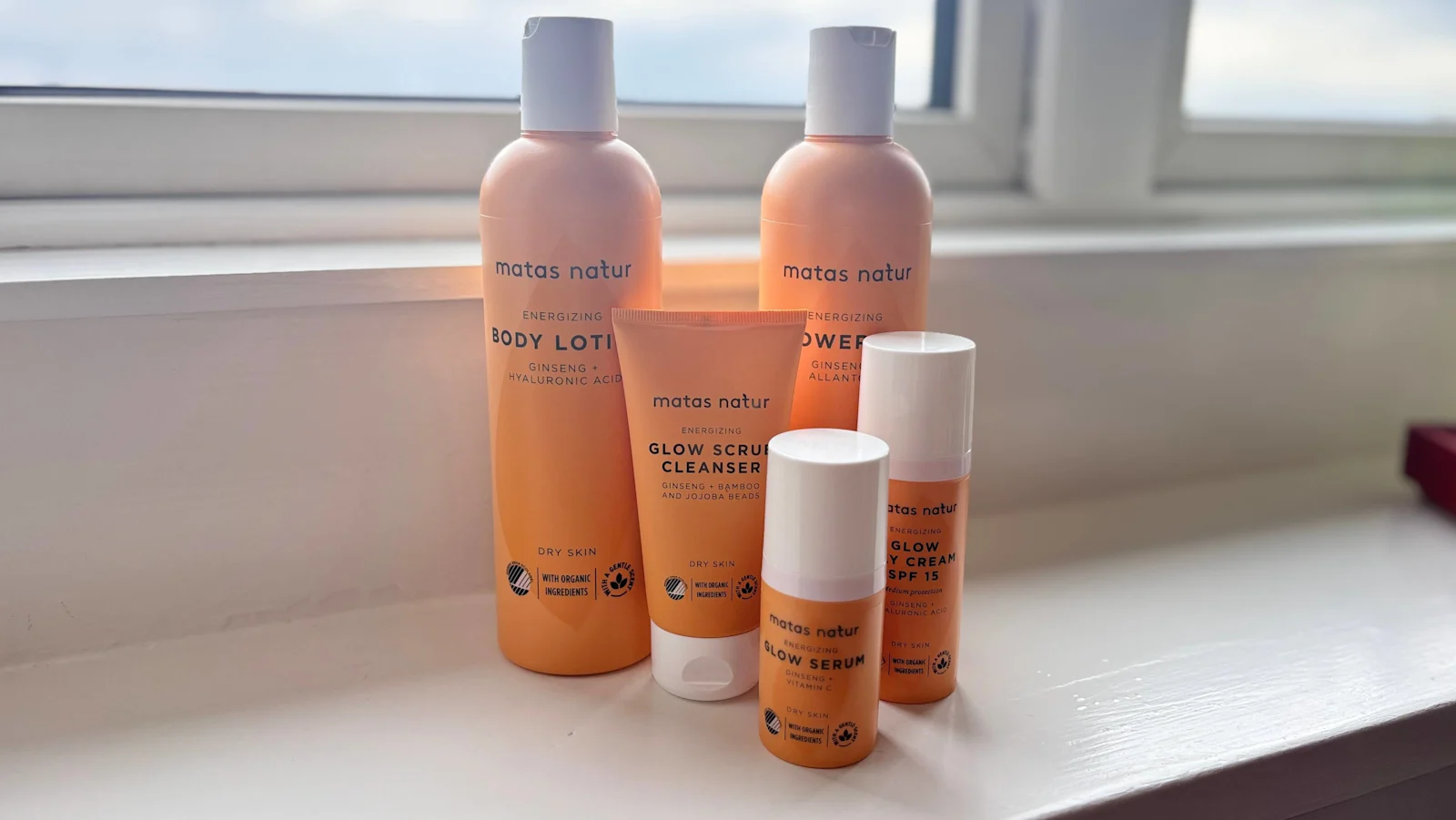 Her ses Matas Natur Glow-serien med Body Lotion, Shower Gel, Glow Scrub Cleanser, Glow Serum og Glow Day Cream SPF 15 på en vindueskarm med dagslys.