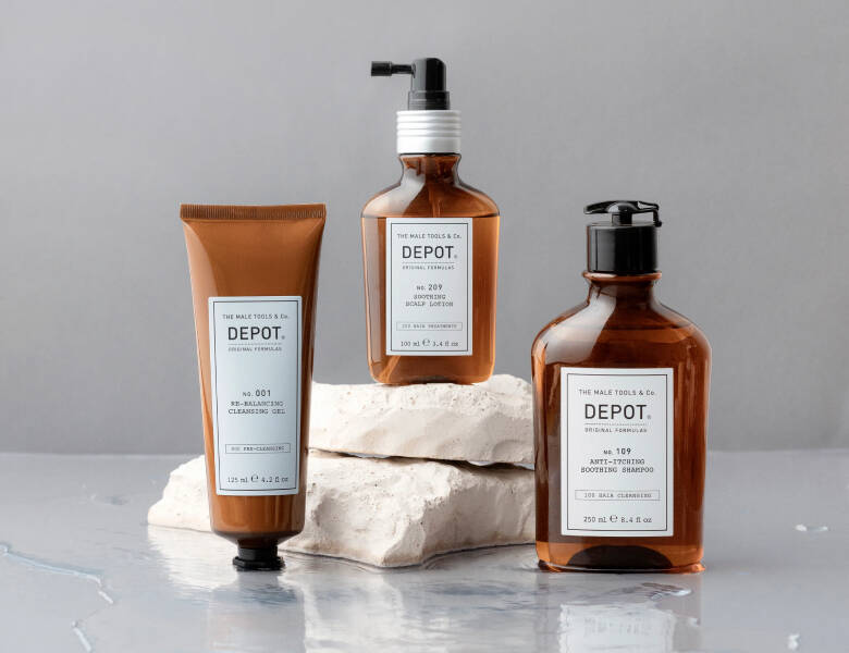 Her ses et billede med tre produkter fra DEPOT placeret på en lys, reflekterende overflade med stenblokke i baggrunden. På produkterne står blandt andet “DEPOT”, “No. 001 Cleansing Gel”, “No. 209 Soothing Scalp Lotion” og “Anti-Dandruff Soothing Shampoo”. Produkterne er i brune emballager med hvide etiketter.