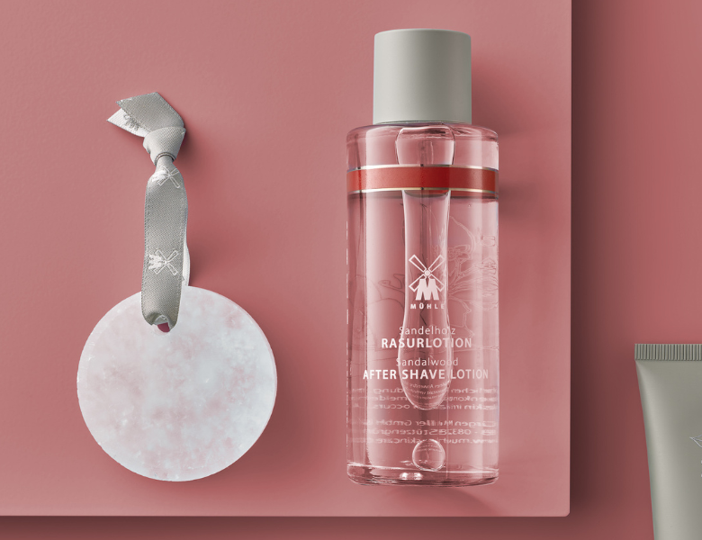 Flaske med Mühle sandalwood after shave lotion står på rosa baggrund ved siden af en rund marmorsten med ophængsbånd.