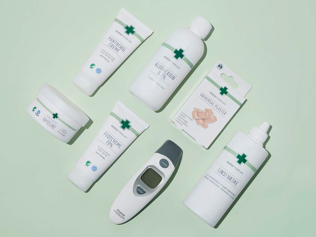 Matas Medicare-produkter ligger fladt på lysegrøn baggrund: panthenolcreme, klorhexidin, universalplastre, vaseline, fedtcreme, linsevask og et digitalt termometer.