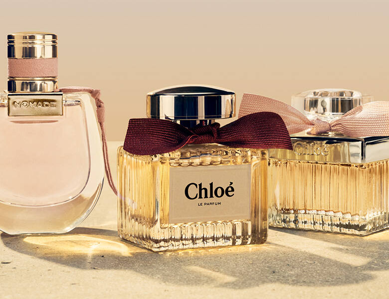 Her ses et billede med tre parfumeflakoner placeret side om side på en lys overflade mod en neutral baggrund. Til venstre ses en flakon med teksten “NOMADE”, og til højre to flakoner med teksten “Chloé”, hvoraf den ene har teksten “LE PARFUM”. Flakonerne er i klare og gyldne nuancer med detaljer som bånd og guldfarvede låg.
