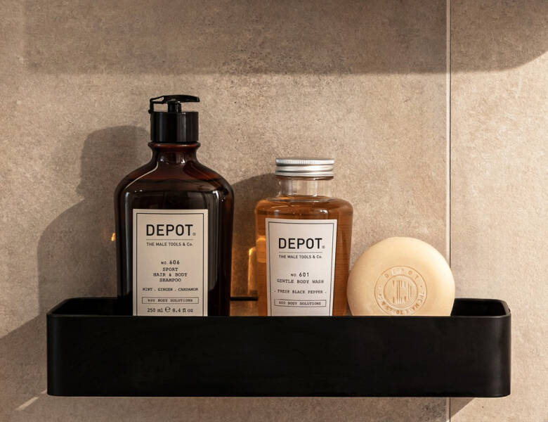 Her ses et billede med tre produkter fra DEPOT placeret på en hylde mod en flisevæg. På produkterne står blandt andet “DEPOT”, “Hair & Body Shampoo” og “Gentle Body Wash”. Produkterne er i brune flasker samt en lys sæbe.