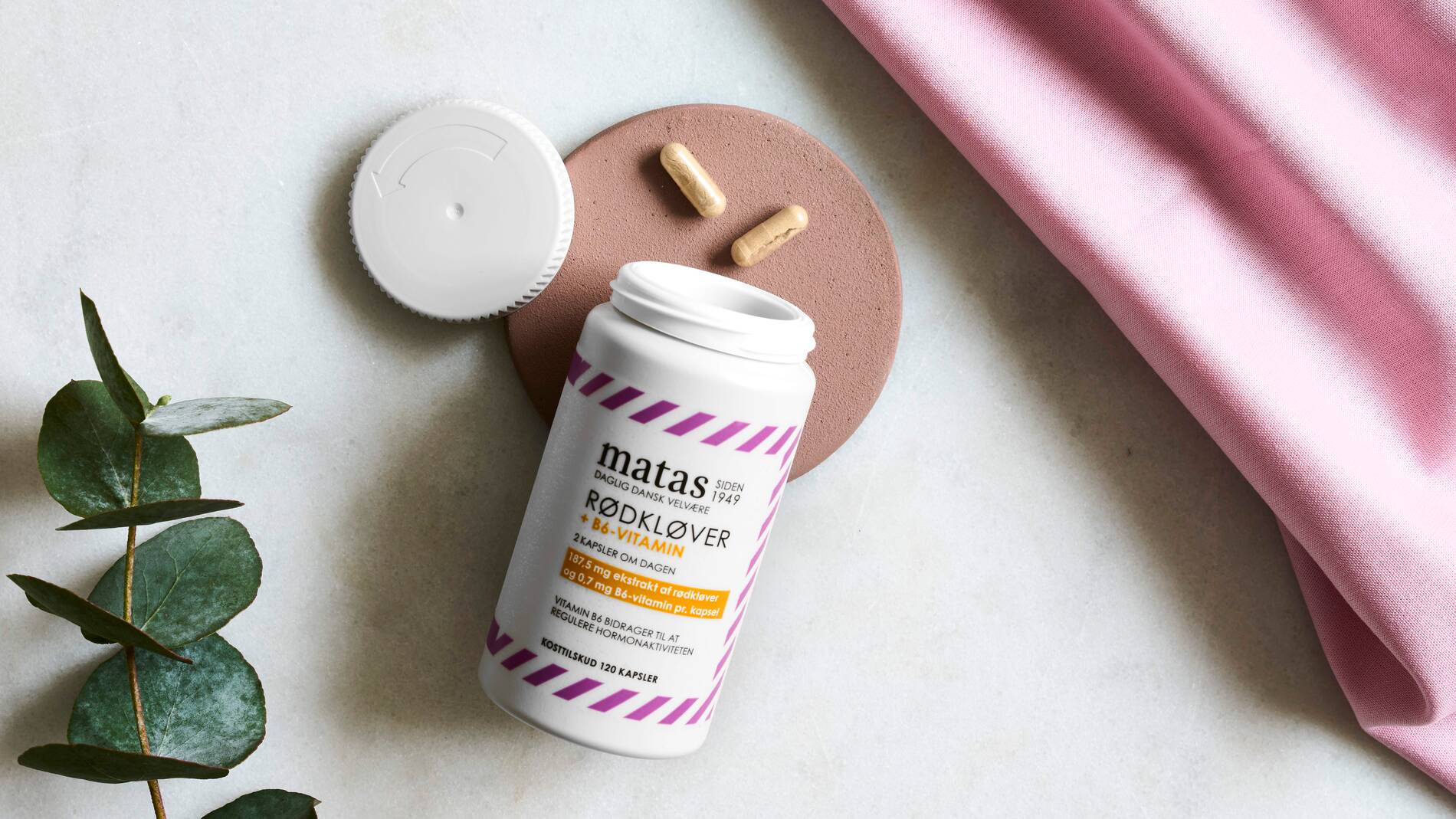 En åben beholder med kosttilskuddet “Matas Rødkløver + B6-vitamin” ligger på en lys overflade sammen med to beige kapsler. Det hvide låg ligger ved siden af beholderen oven på en rund, rosa bakke. Emballagen er hvid med lyserøde striber og sort tekst. I venstre side af billedet ligger en grøn plantekvist, og i højre side ses et stykke lyserødt stof, som giver billedet et blødt og roligt udtryk.