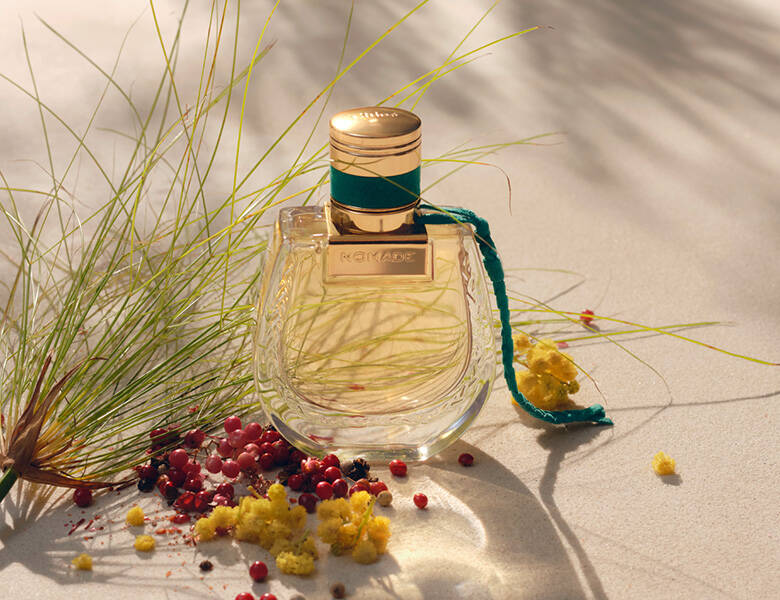 Her ses et billede af en parfumeflakon med teksten “NOMADE” placeret på en lys, sandfarvet overflade. Flakonen er gennemsigtig med gyldne nuancer og har en grøn snor fastgjort ved låget. Omkring flakonen ligger små blomster og bær samt grønne planter.