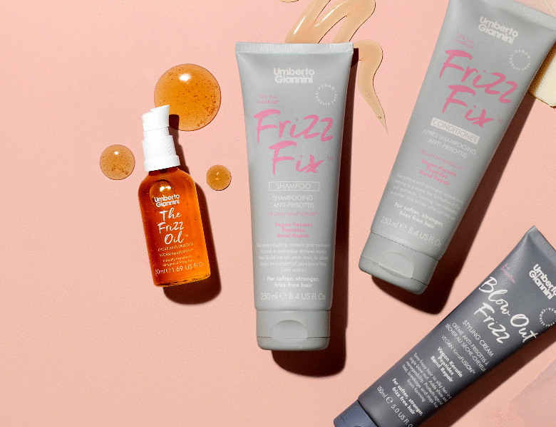 Her ses et billede med flere hårprodukter fra Umberto Giannini placeret mod en lys baggrund. På produkterne står blandt andet “Frizz Fix”, “The Frizz Oil” og “Blow Dry Frizz”. Produkterne er i grå og orange emballage med pink og hvid tekst, og der ses dekorative dråbeelementer omkring dem.
