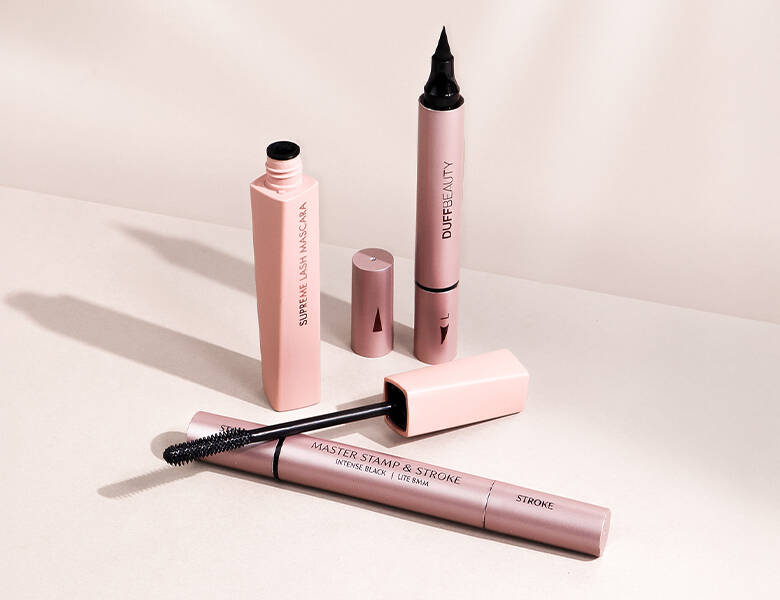 Her ses nogle forskellige makeupprodukter fra DUFFBEAUTY på en lys lyserød baggrund. 
