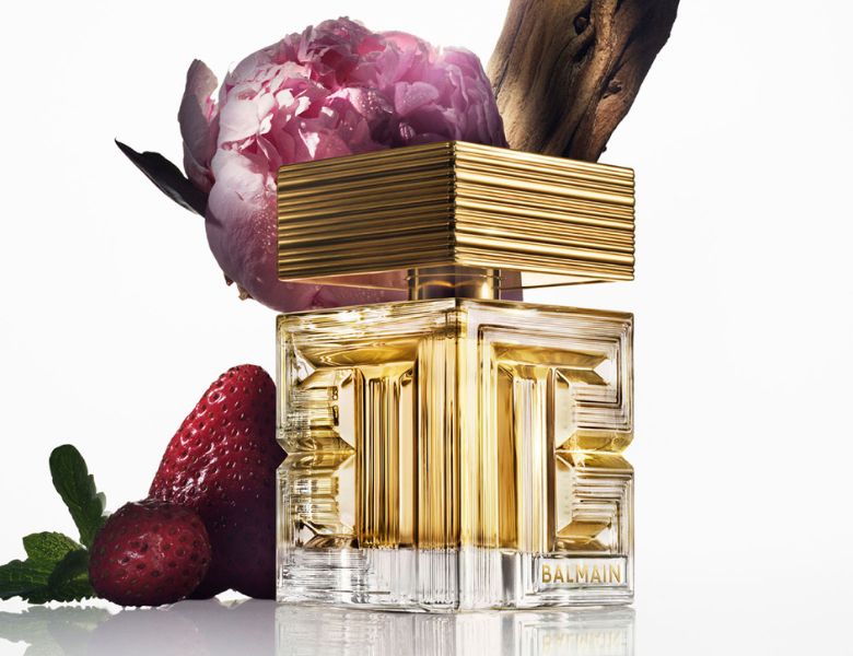 Her ses et billede af en parfumeflakon fra Balmain placeret centralt mod en lys baggrund. Flakonen er i gyldne og transparente nuancer med et geometrisk design. Bag og omkring flakonen ses en lyserød blomst, et stykke træ samt jordbær og grønne blade.