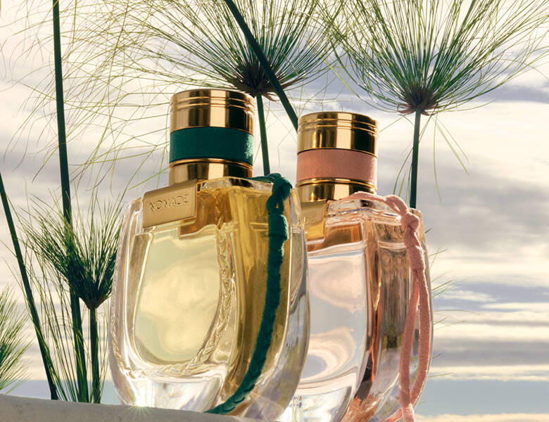 Her ses et billede med to parfumeflakoner med teksten “NOMADE” placeret udendørs mellem høje planter. Flakonerne er gennemsigtige med gyldne nuancer og har henholdsvis en grøn og en rosa snor fastgjort ved halsen. I baggrunden ses himmel og sollys, der skinner gennem planterne.