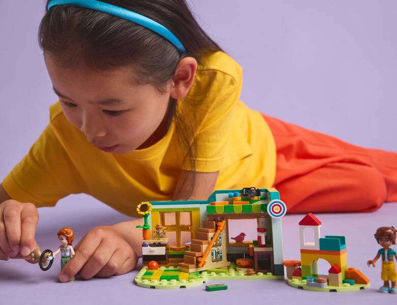 Her ses en lilla baggrund med en pige, som leger med Lego. Pigen har røde bukser, en gul trøje og en blå hårbøjle i sit lange brune hår.