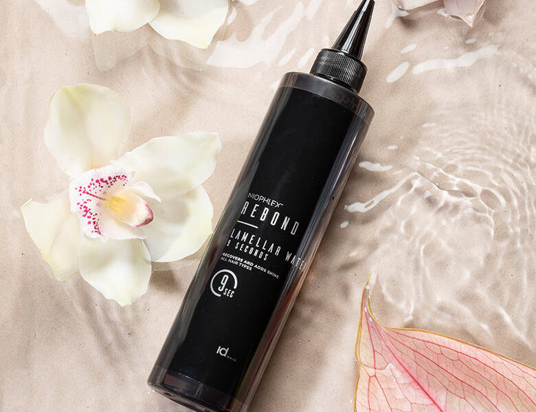 Her ses et sort IdHAIR produkt liggende på en lyserød baggrund med vand og blomster. 