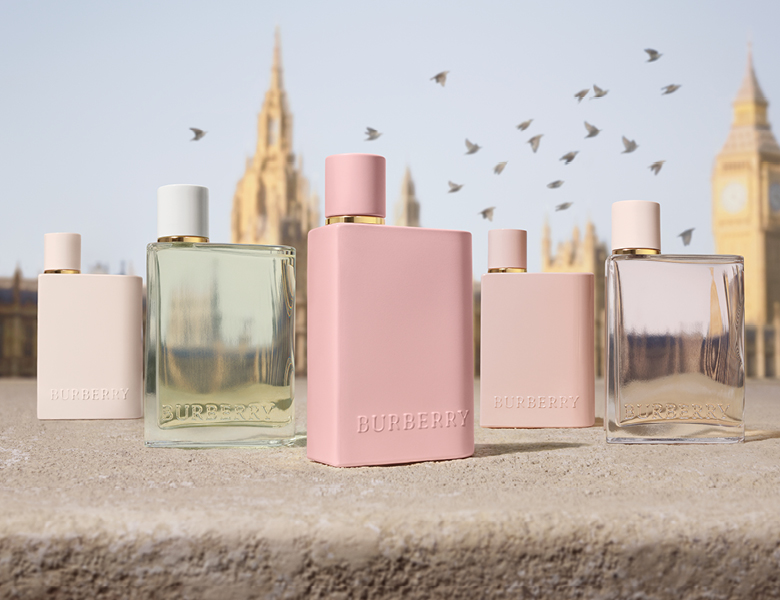 På billedet ses flere Burberry Her-parfumer opstillet foran en sløret baggrund med Big Ben og flyvende fugle.