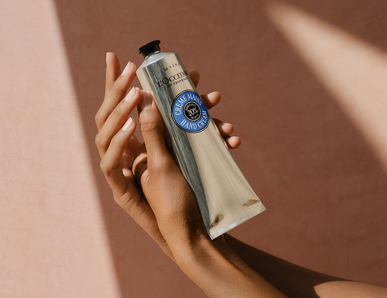 Her ses en hånd, der holder en L’Occitane håndcreme foran en varm beige baggrund.