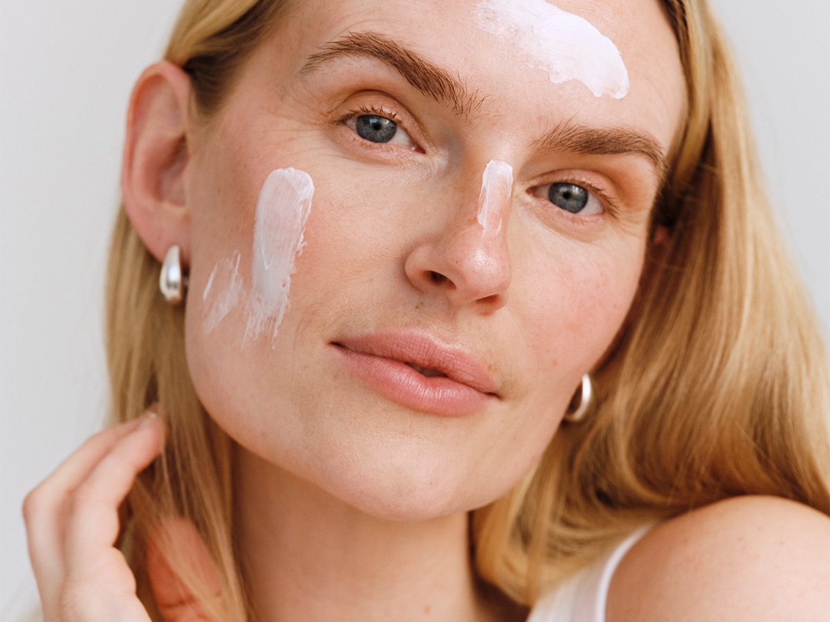 Her ses et nærbillede af en person med lyst hår og sølvfarvede øreringe. Personen har hvid creme påført på panden, næsen og kinden og kigger mod kameraet.