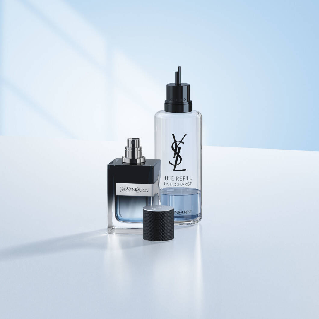 En Yves Saint Laurent Y-parfumeflaske står på en lys overflade foran en blød lyseblå baggrund. Ved siden af står en gennemsigtig refillflaske med sort pumpe og YSL-logo. Parfumeflaskens sorte låg ligger foran produkterne. Billedet har et stilrent og moderne udtryk med fokus på luksusparfume og refill-løsninger.
