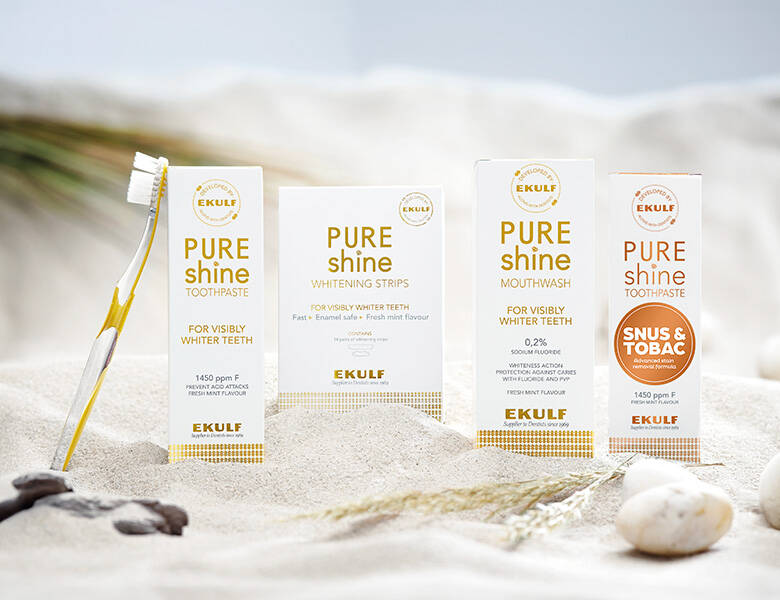 Her ses fire pakker med Ekulf produkter samt en tandbørste i noget sand. Farverne er meget lyse.
