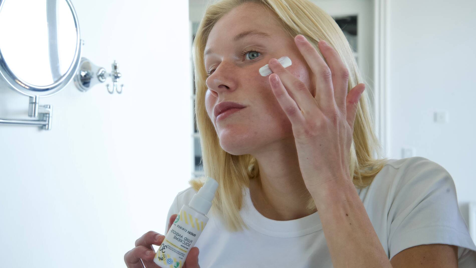 En person med lyst hår påfører ansigtscreme på kinden med fingrene, mens personen holder en flaske med solcreme fra Matas Striber i den anden hånd. Personen står indendørs foran et spejl i et lyst badeværelse og ser fokuseret mod spejlet. Solcremen har hvid emballage med gule striber og flere certificeringsikoner nederst på flasken.