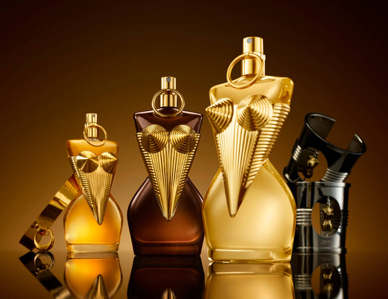 Her ses et billede med flere parfumeflakoner formet som overkroppe placeret side om side. Flakonerne er i forskellige nuancer af guld og mørk brun med detaljer i guldfarve og ringformede låg. Baggrunden er gylden med en glansfuld overflade, hvor produkterne spejles.