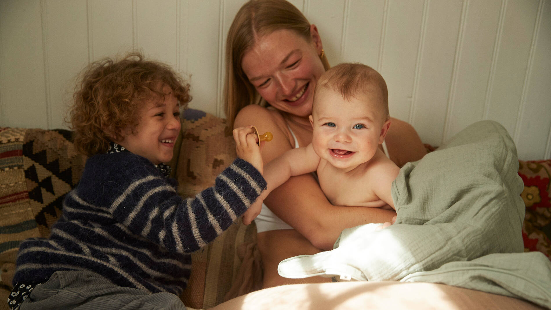En voksen sidder i en sofa og holder en smilende baby i armene, mens et ældre barn med krøllet hår sidder ved siden af og leger med en sut. Alle tre smiler og ser glade ud sammen. Babyen er pakket ind i et lyst tæppe, og scenen er omgivet af bløde puder og tæpper i varme farver. Billedet har en hyggelig og nærværende stemning med varmt sollys, der falder ind i rummet.