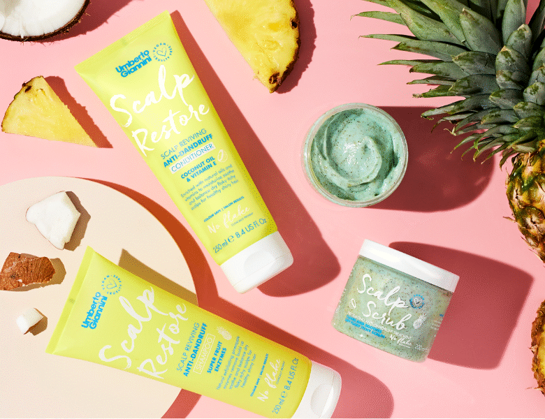 Her ses et billede med hårprodukter fra Umberto Giannini placeret mod en lyserød baggrund. På produkterne står blandt andet “Scalp Restore” og “Scalp Scrub”. Produkterne er i grønne og lyse emballager og er omgivet af ananas og kokosstykker.