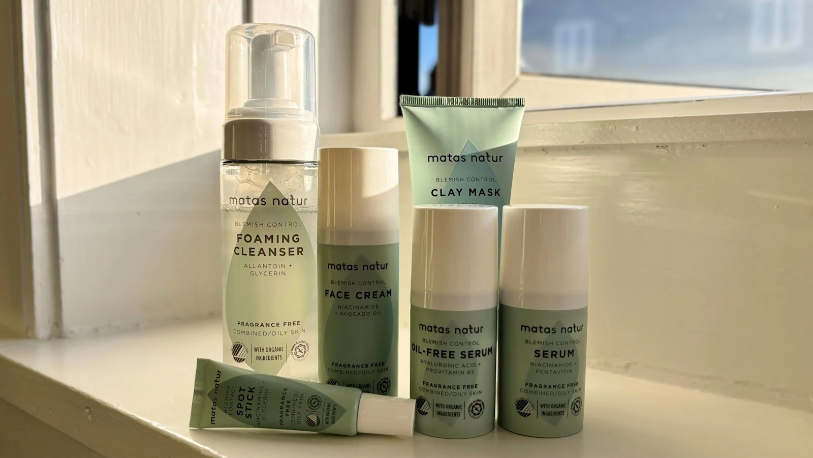 Her ses Matas Natur Blemish Control-serien med Foaming Cleanser, Face Cream, Clay Mask, Oil-Free Serum, Serum og Spot Stick på en vindueskarm i naturligt lys.