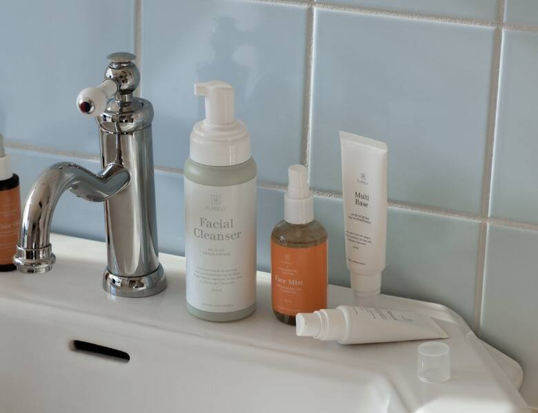 Her ses et billede af flere hudplejeprodukter placeret ved en vask med en sølvfarvet vandhane og lyse fliser i baggrunden. På produkterne står blandt andet “Facial Cleanser”, “Face Mist” og “Multi Base”. Produkterne er i lyse og neutrale emballager.