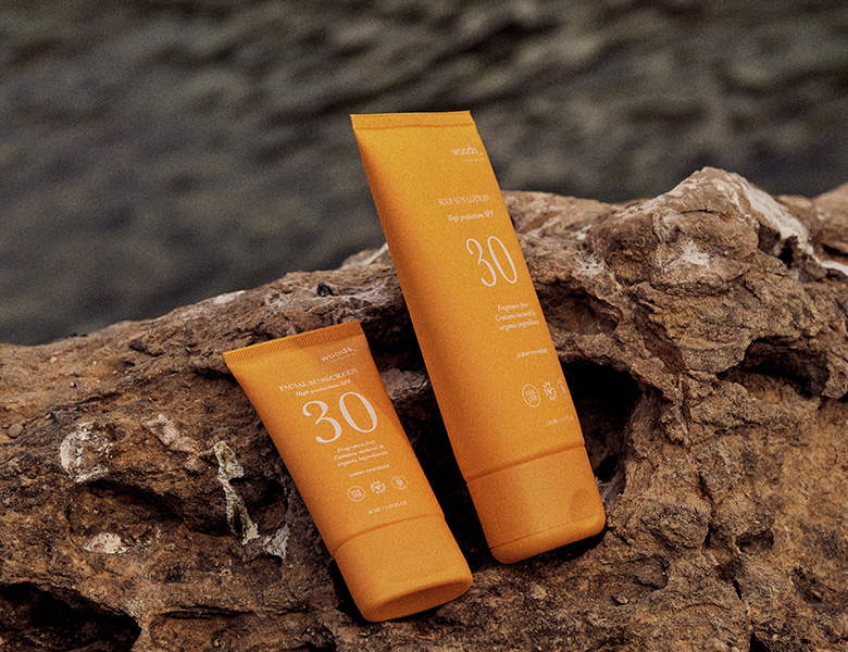 Her ses WOODS_ solcreme i orange emballage placeret på klipper ved vandet.