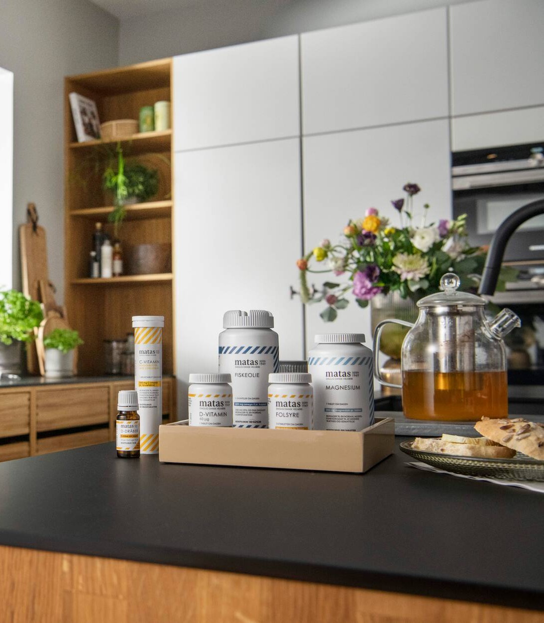 Her ses et udvalg af Matas Striber kosttilskud, herunder fiskeolie, magnesium, folsyre, D-vitamin, D-dråber og C-vitamin, samlet på en bakke i et køkken.