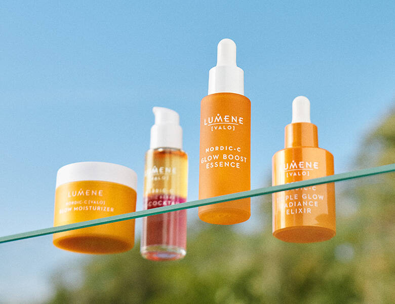 Her ses fire forskellige orange Lumene produkter. Baggrunden er nogle grønne træer og en blå skyfri himmel.