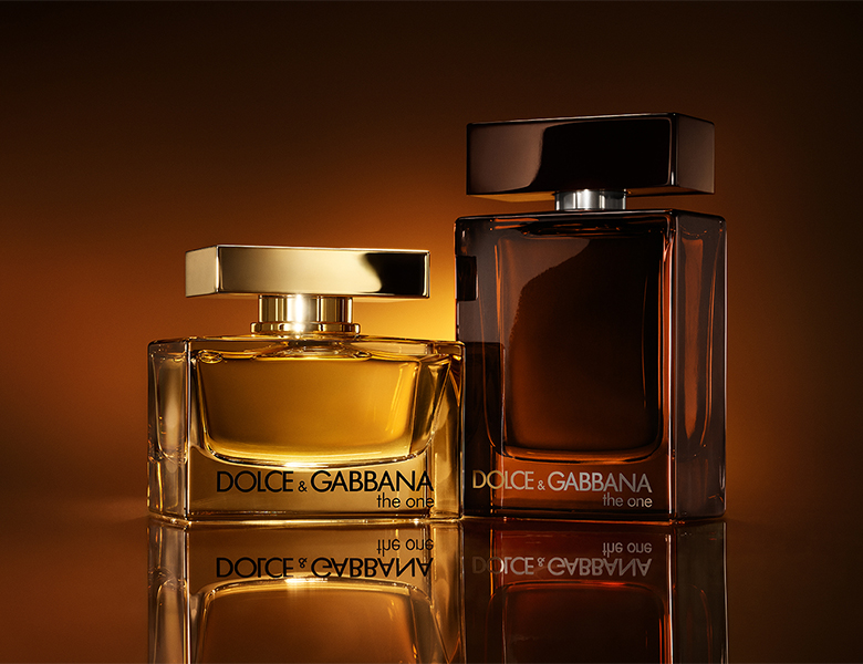 Her ses et billede med to parfumeflakoner fra Dolce & Gabbana placeret side om side mod en mørk baggrund med gyldent lys. På flakonerne står teksten “DOLCE & GABBANA the one”. Flakonerne er i gyldne og mørke nuancer med firkantede låg.