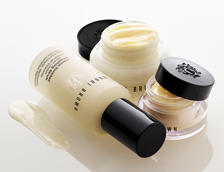 Her ses et billede med hudplejeprodukter fra Bobbi Brown mod en lys baggrund. Produkterne omfatter blandt andet en Vitamin Enriched Face Base, en Vitamin Enriched Face Base Eye Cream og en lotion. Krukkerne er åbne, så den cremede tekstur er synlig, og en lille mængde creme er påført ved siden af produkterne for at fremhæve konsistensen.