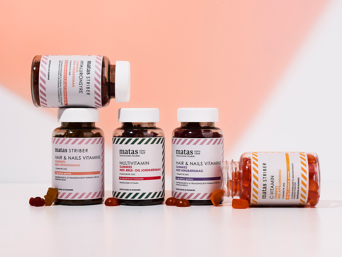 Her ses et udvalg af Matas Striber vitamin-gummies i brune beholdere med hvide låg. Sortimentet omfatter Hair & Nails Vitamins, Multivitamin, Hyaluronsyre og C-vitamin, og flere farvede gummies ligger foran produkterne mod en rosa og hvid baggrund.