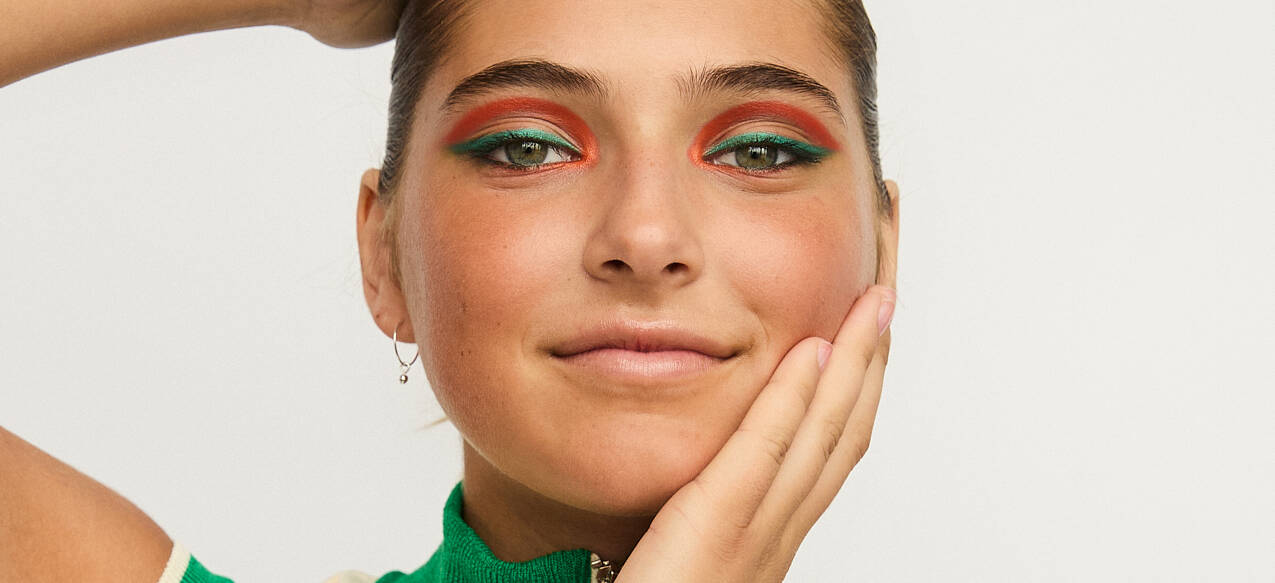 Her ses et billede af en person mod en lys baggrund med farverig øjenmakeup i grønne og orange nuancer. Personen holder en hånd mod kinden og en arm over hovedet.