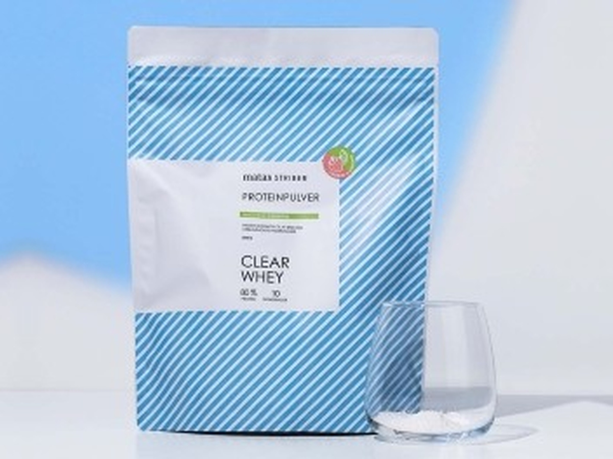 En pose Matas Striber Clear Whey proteinpulver står ved siden af et glas med hvidt pulver på lyseblå baggrund.
