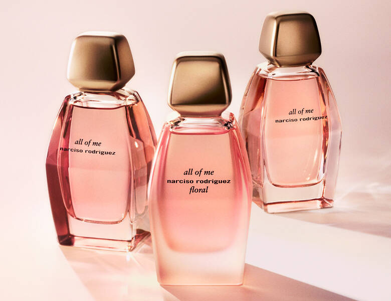 Her ses et billede med tre parfumeflakoner i rosa nuancer placeret side om side mod en lys baggrund. På flakonerne står teksten “all of me narciso rodriguez”, og på den midterste står også “floral”. Flakonerne har kantede former med gyldne låg.