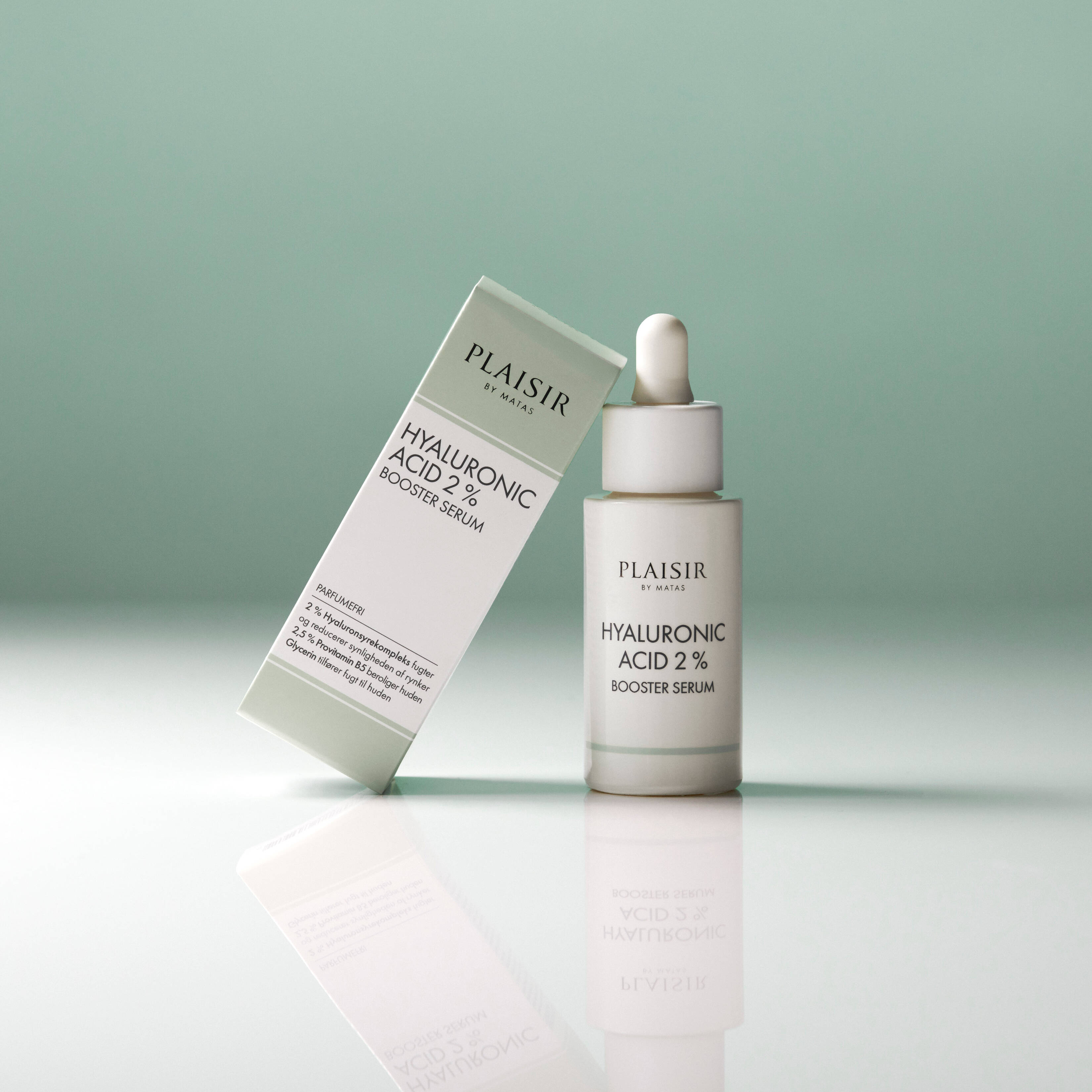 En flaske Plaisir Hyaluronic Acid 2% Booster Serum står på en blank overflade mod en grøn baggrund. Ved siden af står produktets æske let lænet. Flasken har pipette og spejler sig i underlaget.