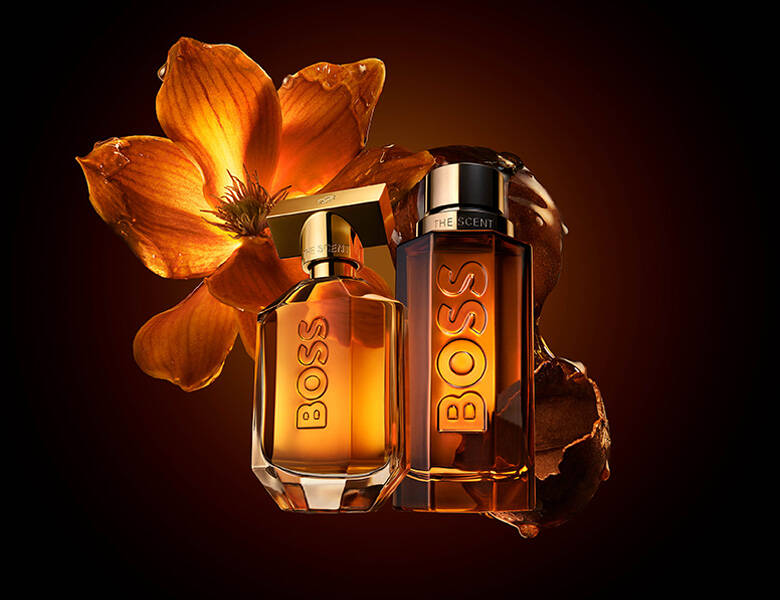 Her ses et billede med to parfumeflakoner fra BOSS placeret side om side mod en mørk baggrund med orange lys. På flakonerne står teksten “BOSS”, og på den ene også “THE SCENT”. Bag produkterne ses en orange blomst og mørke frugtelementer.