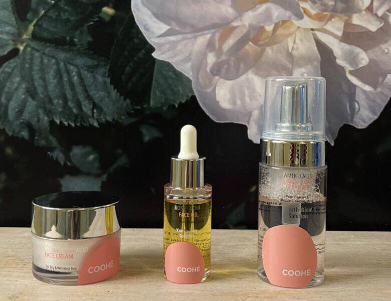 Tre Coohe hudplejeprodukter står på en lys overflade foran mørk blomsterbaggrund – creme i krukke, ansigtsolie med pipette og serum i pumpeflaske.