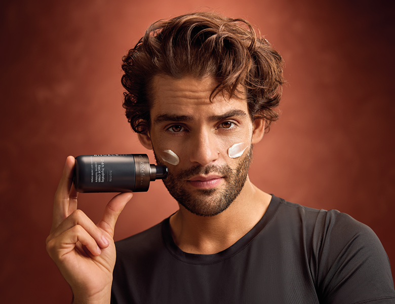 Her ses en mand med creme i ansigtet, der holder et RITUALS hudplejeprodukt foran en varm brun baggrund.