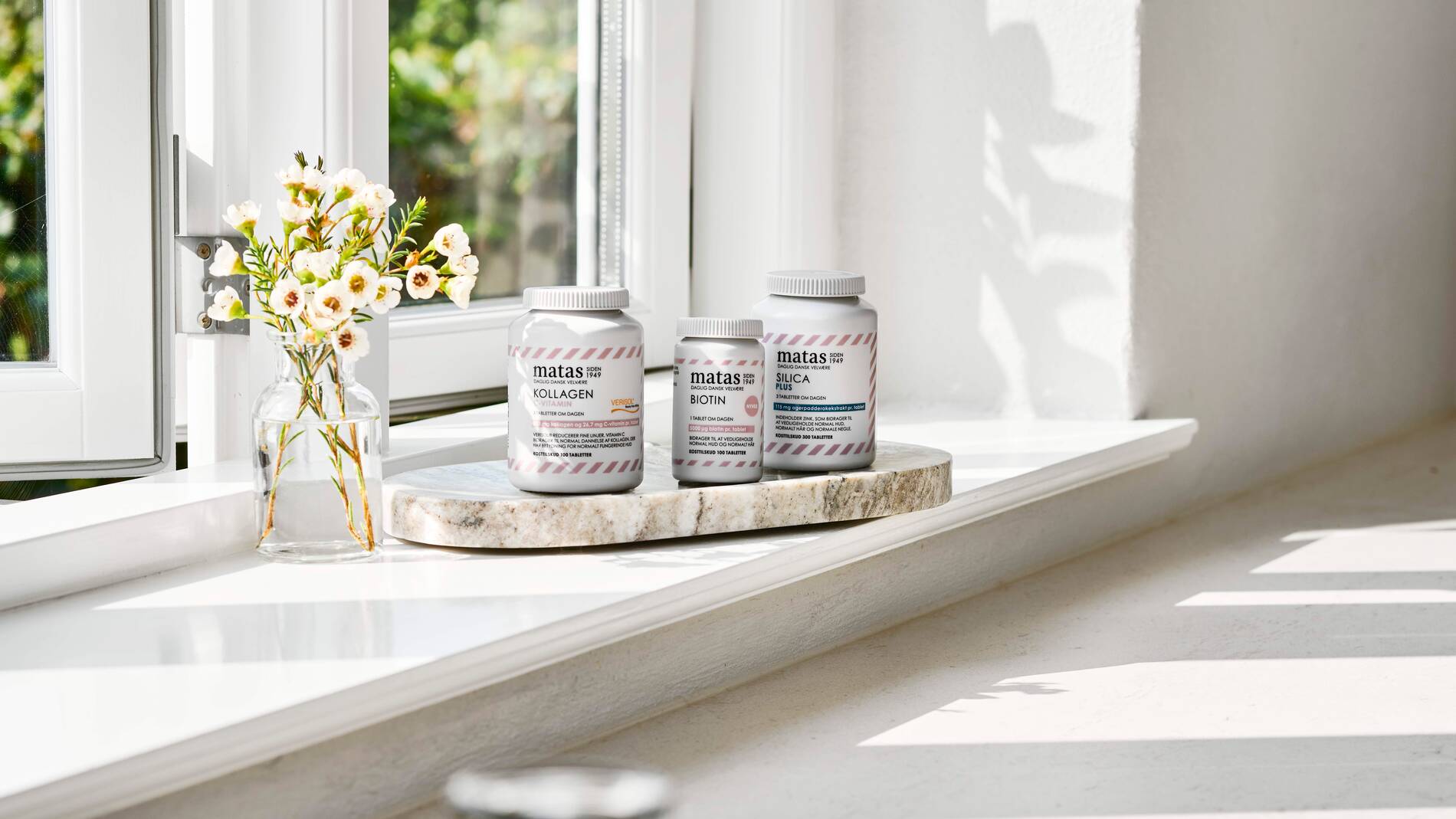 Her ses Matas Striber Kollagen, Biotin og Silica Plus på et marmorfad i en vindueskarm ved siden af en vase med blomster.