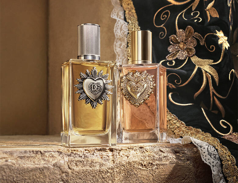 To Dolce & Gabbana parfumer i klare flasker med dekorative hjerteemblemer står på sten foran broderet stof i sort og guld
