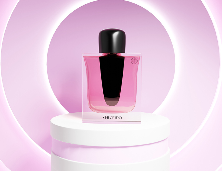 Her ses et billede af en parfumeflakon fra Shiseido placeret på et podium mod en pink baggrund med cirkulært lys. Flakonen er i pink nuance med sort låg og har teksten “SHISEIDO” nederst på flasken.