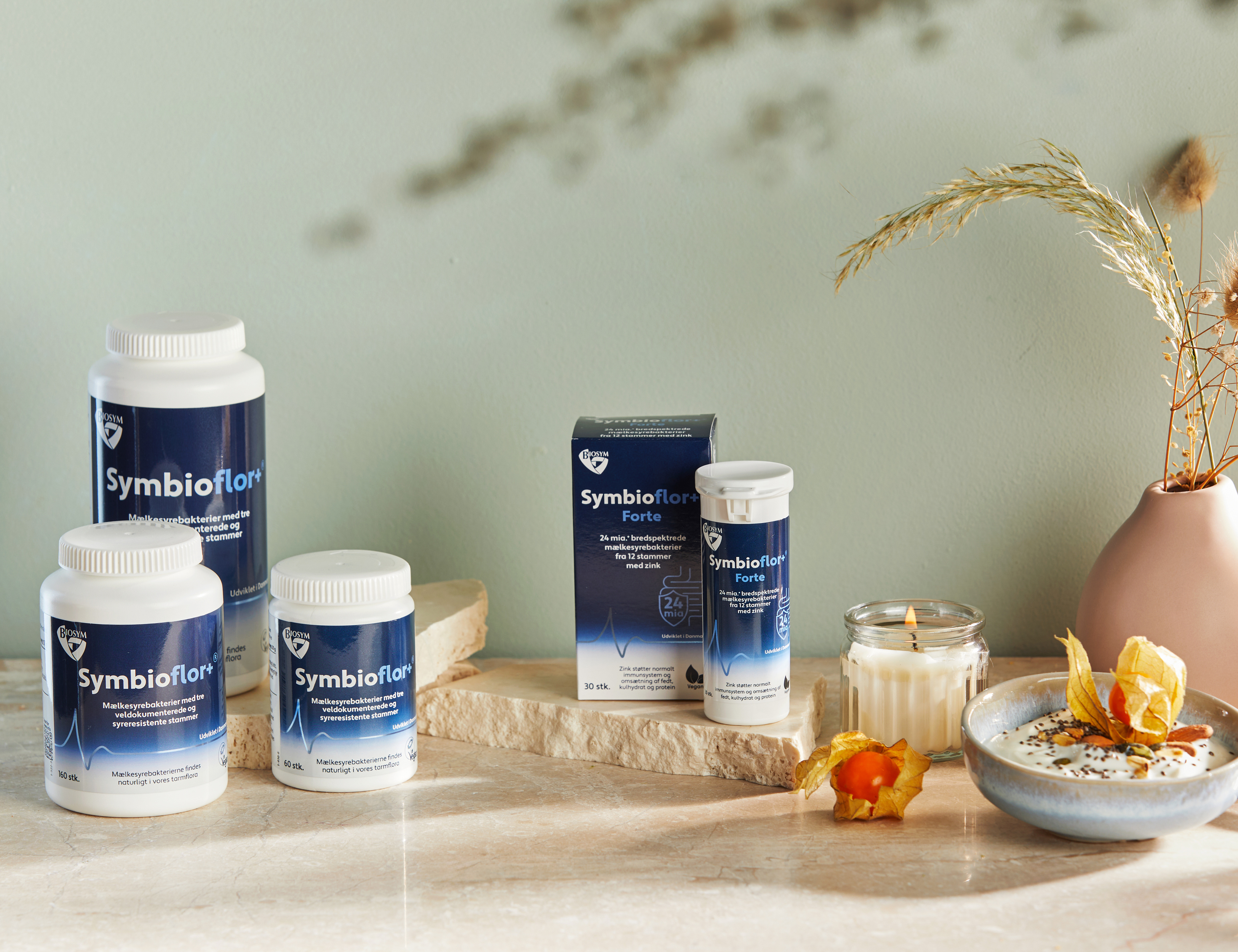 Symbioflor kosttilskud står på lyse stenplader foran en lys grøn baggrund ved siden af et tændt lys, en vase med tørrede blomster og en skål med yoghurt og frugt.