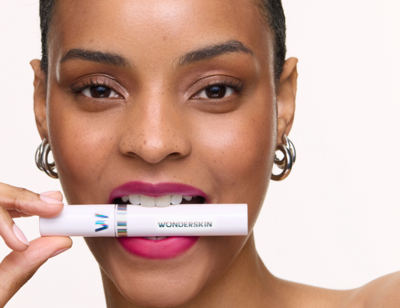Her ses et nærbillede af en smilende person med en intens pink læbefarve, der holder et Wonderskin Wonder Blading Lip Stain vandret foran munden. Fokus er på læbeproduktet og det færdige resultat på læberne.