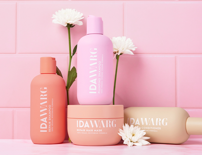 Her ses IDA WARG hårplejeprodukter i lyserød og beige emballage sammen med blomster på en lyserød baggrund.
