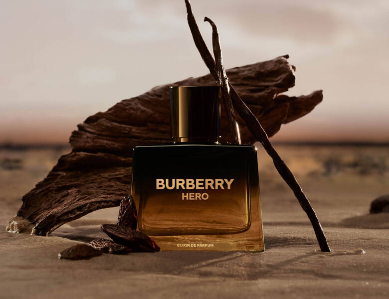 Her ses et billede af en parfumeflakon fra Burberry placeret på en sandet overflade foran et stykke drivtømmer. På flakonen står teksten “BURBERRY HERO” samt “ELIXIR DE PARFUM”. Ved siden af flakonen ses vaniljestænger.