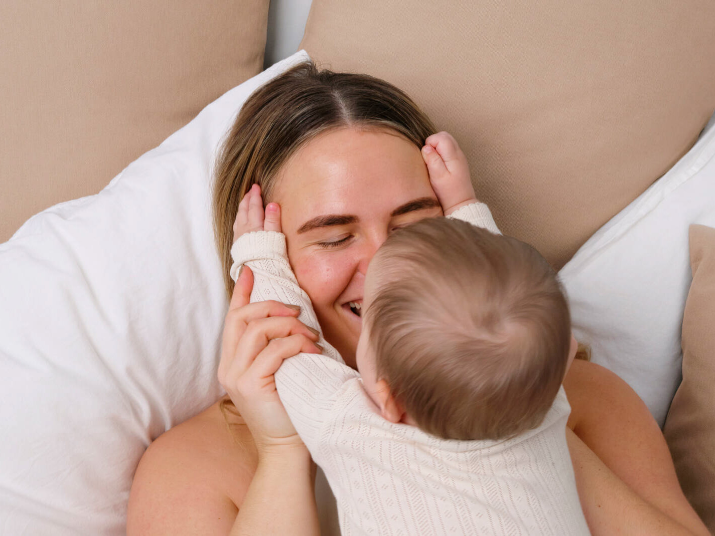 Mor og baby ligger tæt i en seng med hvide dyner og beige puder, mens babyen holder om morens ansigt og de smiler til hinanden.
