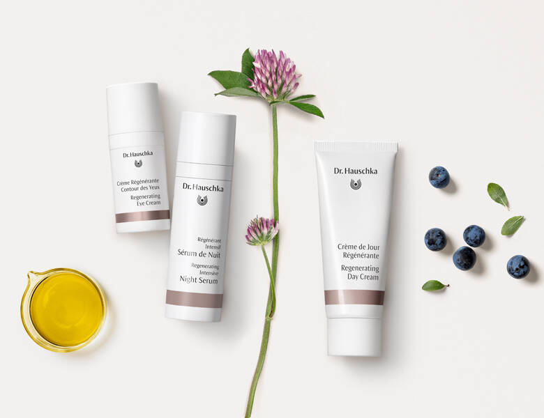 Her ses en hvid baggrund med tre produkter, som alle har en lilla stribe. Der ses desuden en gul væske, en lilla blomst og nogle blåbær. 