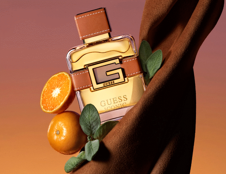 Her ses et billede af en parfumeflakon med teksten “GUESS ICONIC” placeret mod en varm, orange baggrund. Flakonen har guldfarvede detaljer og en brun rem med et “G”-logo. Omkring flakonen ses appelsiner og grønne blade samt et stykke stof.